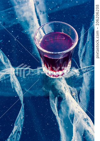 Alcohol in a glass and clean ice with beautiful deep cracks. A glass with red tincture stands on the transparent ice of the lake. Vertical. 106206208
