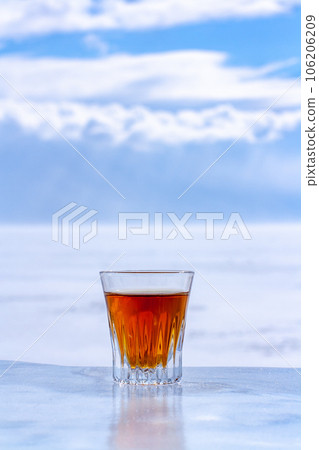 Whiskey in a glass stands on an ice surface on a blurry light background. Orange alcoholic drink in a glass. Copy space. Side view. Vertical. 106206209