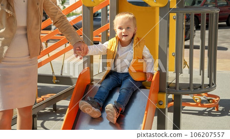 Cute baby boy holding mothers hand while riding down the slide on playground. Happy parenting, family having time together, kids and parents outdoors. 106207557