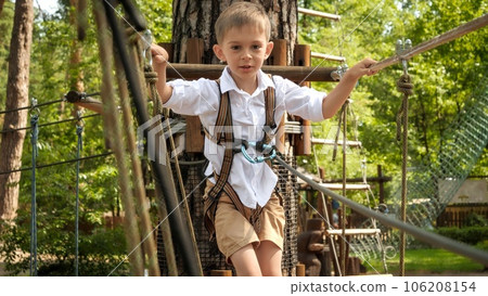 Portrait of little boy having fun while crossing rope bridge in summer camp. Active childhood, healthy lifestyle, kids playing outdoors, children in nature. 106208154