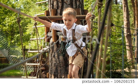 Happy smiling boy having fun climbing and crossing obstacles in rope park. Active childhood, healthy lifestyle, kids playing outdoors, children in nature. Happy smiling boy having fun climbing and crossing obstacles in rope park. Active childhood, healthy lifestyle, kids playing outdoors, children in nature. 106208155