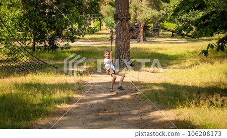 Happy laughing boy having fun riding on the zip line at summer park. Active childhood, healthy lifestyle, kids playing outdoors, children in nature. 106208173