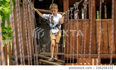 Little boy tied to safety rope crossing wobbly rope bridge at summer adventure park. Kids sports, summer holiday, fun outdoors, scouts. 106208191
