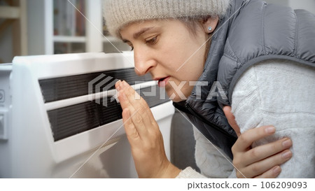 Portrait of woman feeling cold in her house sitting at electric heater. Concept of energy crisis, high bills, economy and saving money on monthly utility payments. 106209093