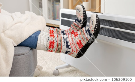 Feet in warm socks warming under blanket at electric heater. Concept of energy crisis, high bills, economy and saving money on monthly utility payments. Feet in warm socks warming under blanket at electric heater. Concept of energy crisis, high bills, economy and saving money on monthly utility payments. 106209100
