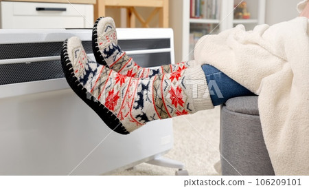 Closeup of woman wearing warm socks sitting at electric heater in living room. Concept of energy crisis, high bills, economy and saving money on monthly utility payments. Closeup of woman wearing warm socks sitting at electric heater in living room. Concept of energy crisis, high bills, economy and saving money on monthly utility payments. 106209101