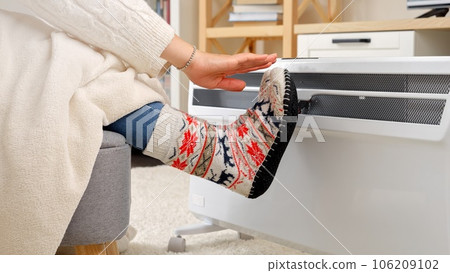 Closeup of woman warming hands and feet in woolen socks at electric heater. Concept of energy crisis, high bills, economy and saving money on monthly utility payments. Closeup of woman warming hands and feet in woolen socks at electric heater. Concept of energy crisis, high bills, economy and saving money on monthly utility payments. 106209102