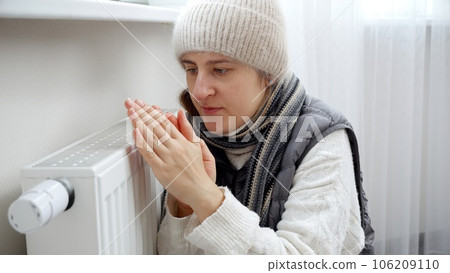 Young woman feeling cold in house sitting at heating radiator. Concept of energy crisis, high bills, broken heating system, economy and saving money on monthly utility payments. 106209110