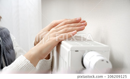 Closeup of young woman in sweater warming her hands at heater in cold home 106209125