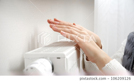 Woman in sweater and scarf warming her hands at heating radiator on the cold day. Concept of energy crisis, high bills, broken heating system, economy and saving money on monthly utility payments. 106209126
