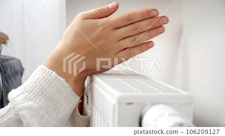 Closeup of woman warming her hands at heating radiator in house. Concept of energy crisis, high bills, broken heating system, economy and saving money on monthly utility payments. 106209127