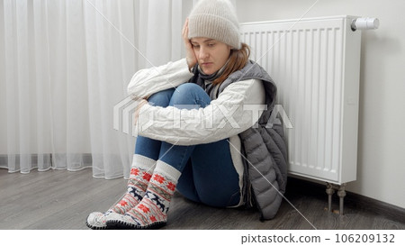 Young woman in warm clothes sitting at broken heating radiator in house. Concept of energy crisis, high bills, broken heating system, economy and saving money on monthly utility payments. 106209132
