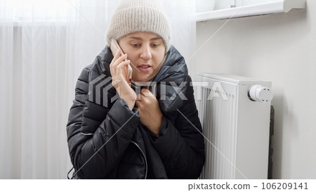 Young woman in winter coat and hat sitting by the broken heater in freezing home and calling repair service 106209141