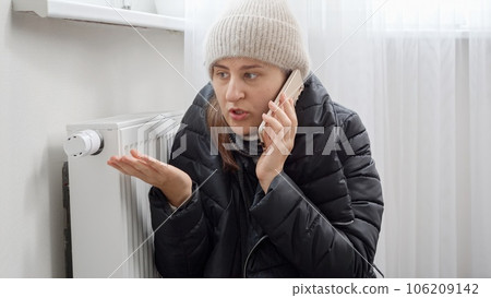 Woman in coat and hat at home calling repairman to fix heating system at house. Concept of energy crisis, high bills, broken heating system, economy and saving money on monthly utility payments 106209142