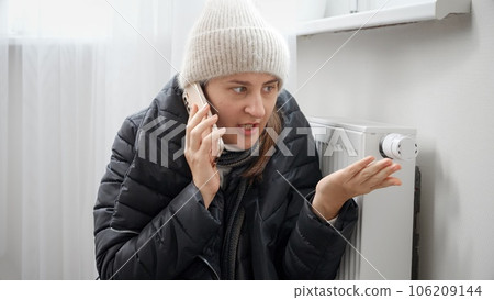 Woman freezing at home talking with repairman by phone. Concept of energy crisis, high bills, broken heating system, economy and saving money on monthly utility payments. 106209144