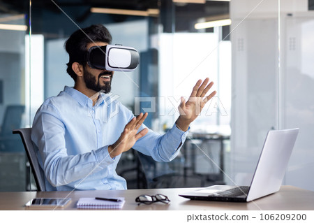 Young Indian male student sitting at a desk in the office wearing a virtual mask, watching online videos, studying, talking, gesturing with his hands. 106209200