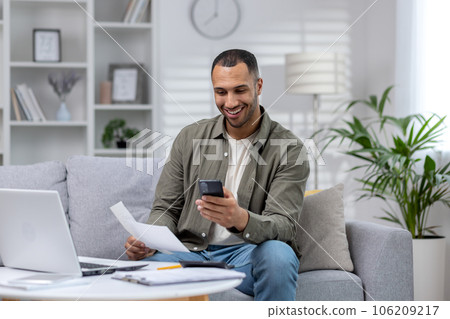 Young smiling African-American man sitting on sofa at home, holding phone and documents, working on laptop. Young smiling African-American man sitting on sofa at home, holding phone and documents, working on laptop. 106209217