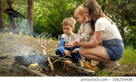 Young mother with two sons cooking sausages on bonfire in camp. Active leisure, children in camping, family vacation in nature. Young mother with two sons cooking sausages on bonfire in camp. Active leisure, children in camping, family vacation in nature. 106209486