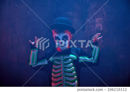 Little boy, child with creepy pale face makeup, wearing costume of skeleton against dark background in neon light with smoke 106210112