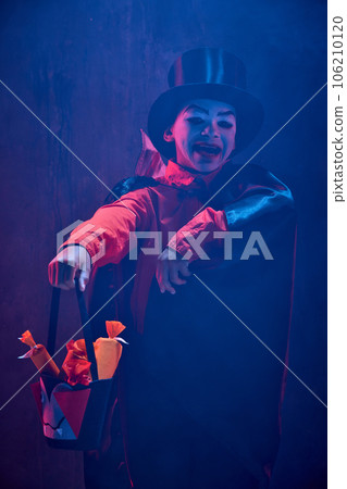 Little boy, child in image of creepy vampire costume and makeup standing with basket for sweets against dark background in neon light with smoke 106210120