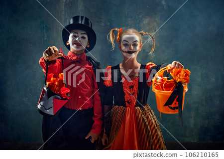 Trick or treat. Boy and girl, children wearing costumes for halloween, standing with basket for candies against dark vintage background Trick or treat. Boy and girl, children wearing costumes for halloween, standing with basket for candies against dark vintage background 106210165