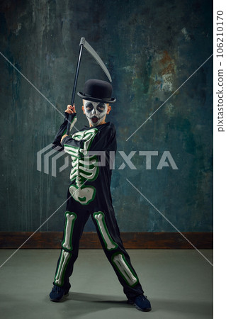 Fantasy. Little boy, child with pale face makeup, wearing costume of skeleton, standing with scythe against vintage green background 106210170