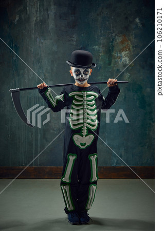 Little boy, child in creative costume with skeleton image and creepy makeup standing with scythe against dark vintage background. 106210171