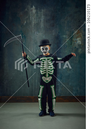 Fantasy. Little boy, child with pale face makeup, wearing costume of skeleton, standing with scythe against vintage green background 106210173