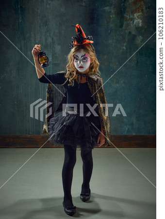 Little girl, child in image of witch, with creepy makeup and costume standing against dark vintage background. Spooky season 106210183