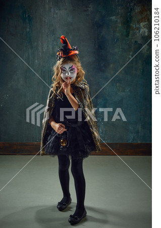 Little girl, child with witch makeup and costume standing against dark vintage background. Mystery 106210184
