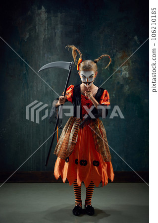 Shh. Keeping secret. Little girl with creepy makeup, costume for halloween standing with scythe against vintage green background 106210185