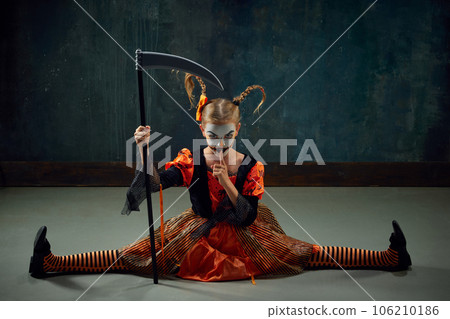Shh. Keeping secret. Fearful little girl with creepy makeup, costume for halloween sitting with scythe against vintage green background 106210186