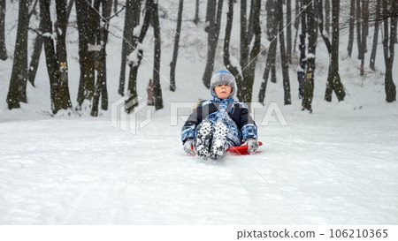Boy gleefully gliding down a snow-covered hill on his plastic sled, grinning from ear to ear. The perfect image of winter holidays, fun in the snow, and the joy of childhood. 106210365