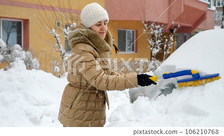 Young woman brushing off snow from her car on a snowy parking lot. Concept of winter, maintenance, and transportation. Young woman brushing off snow from her car on a snowy parking lot. Concept of winter, maintenance, and transportation. 106210768