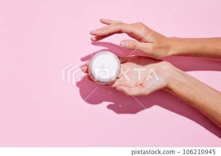 Cropped top view of female hands holding container with cream in hands. Using cosmetics product isolated pastel pink background. Cropped top view of female hands holding container with cream in hands. Using cosmetics product isolated pastel pink background. 106210945