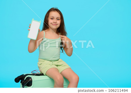Adorable little traveler girl on a plastic blue valise, showing at camera a smartphone with copy space for mobile app Adorable little traveler girl on a plastic blue valise, showing at camera a smartphone with copy space for mobile app 106211494