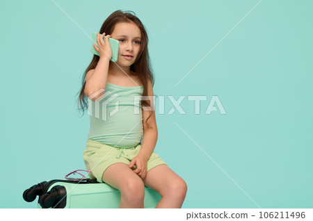 Adorable little kid girl talks on mphone, sitting on suitcase, awaiting flight, checking mobile app, isolated background Adorable little kid girl talks on mphone, sitting on suitcase, awaiting flight, checking mobile app, isolated background 106211496