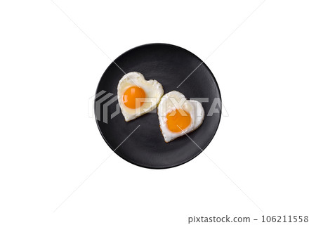 Two heart-shaped fried eggs on a black ceramic plate on a dark concrete background 106211558