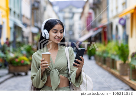 Beautiful young Indian woman in headphones stands on the street with a phone, listens to music, walks, drinks coffee. Beautiful young Indian woman in headphones stands on the street with a phone, listens to music, walks, drinks coffee. 106211858