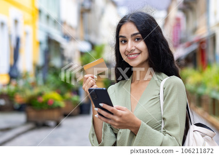 A portrait of a young beautiful Indian woman standing on the street, holding a crew and a phone in her hands, smiles at the camera. The photo is close. A portrait of a young beautiful Indian woman standing on the street, holding a crew and a phone in her hands, smiles at the camera. The photo is close. 106211872
