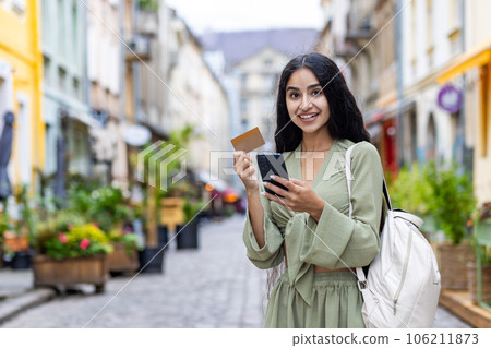 Beautiful young Indian woman using phone and credit card. Standing outside on a city street and smiling at the camera. 106211873