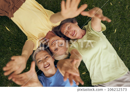 Smiling mature women friends lying head to head on grass and extend their hands to the camera 106212573