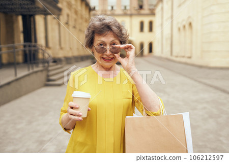 Happy mature woman in sunglasses walking with her shopping bags and coffee cup in city street Happy mature woman in sunglasses walking with her shopping bags and coffee cup in city street 106212597
