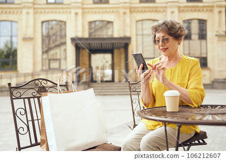 Smiling senior woman is using smartphone while sitting at cafe terrace with coffee after shopping Smiling senior woman is using smartphone while sitting at cafe terrace with coffee after shopping 106212607