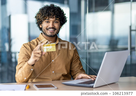 Portrait of young successful hispanic businessman inside office, man smiling and looking at camera, entrepreneur holding bank credit card in hands, satisfied with results of money transfer. 106213977