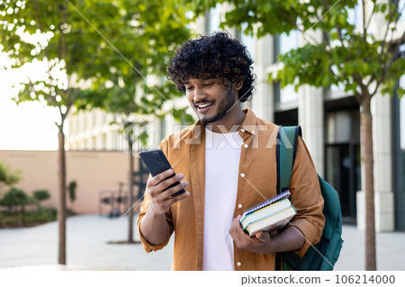 Young smiling Indian male student came to study, standing outside campus, university and using phone. He was holding books and a backpack. 106214000