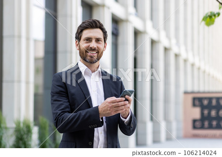 Young business man, office worker standing outside on the street in a suit and using a mobile phone. Smiling at the camera. 106214024