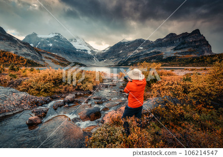 Travel photographer man taking a photo with camera at mount assiniboine in autumn wilderness by lake magog on moody day Travel photographer man taking a photo with camera at mount assiniboine in autumn wilderness by lake magog on moody day 106214547