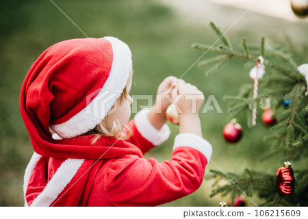 Christmas in july. Child waiting for Christmas in wood in summer. portrait of little girl in red dress decorating christmas tree. winter holidays and people concept. Merry Christmas and Happy Holidays Christmas in july. Child waiting for Christmas in wood in summer. portrait of little girl in red dress decorating christmas tree. winter holidays and people concept. Merry Christmas and Happy Holidays 106215609