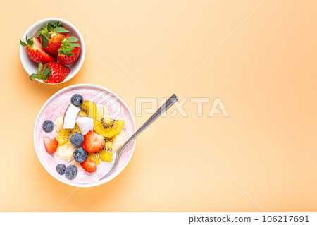 Healthy breakfast or dessert yogurt bowl with fresh banana, strawberry, blueberry, cocos, kiwi top view on minimal pastel paper background and spoon. Space for text 106217691